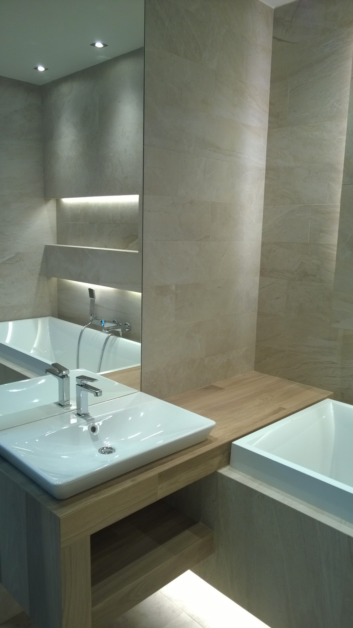 Nowoczesna łazienka z beżowymi płytkami imitującymi kamień, białą prostokątną umywalką nablatową na drewnianej szafce z półką, oraz wanną obudowaną płytkami w tym samym odcieniu, oświetlenie LED...