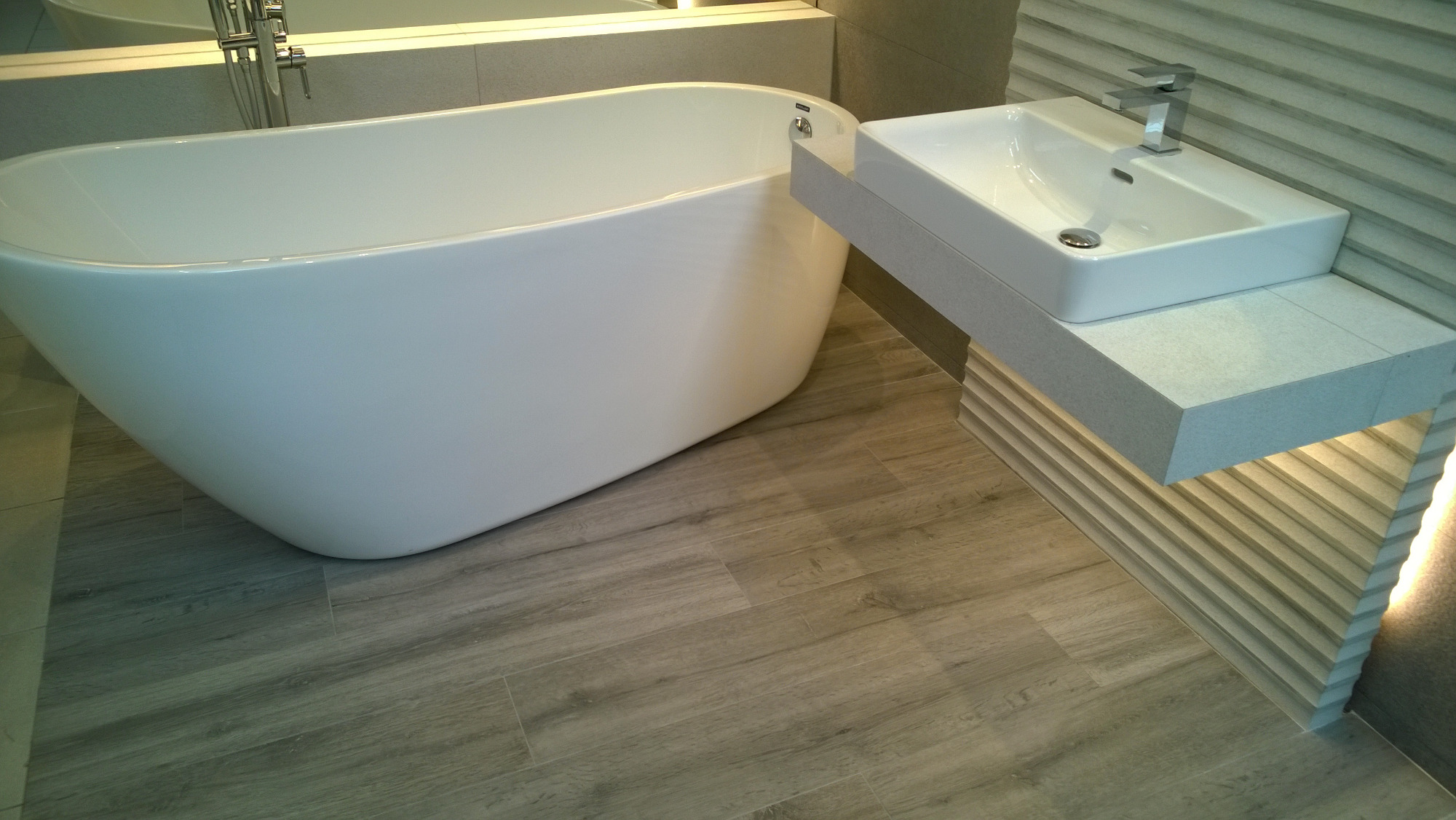 Nowoczesna łazienka z wolnostojącą białą wanną, prostokątną umywalką nablatową i podświetlanym panelem ściennym, wykończona płytkami imitującymi drewno.