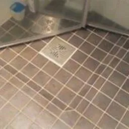 Ułożona pod kątem, ciemna, kwadratowa podłoga w kabinie prysznicowej z widocznym odpływem liniowym i fragmentem szklanych drzwi kabiny.