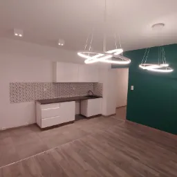 Nowoczesne wnętrze z białymi szafkami kuchennymi, geometryczną mozaiką nad blatem, ciemnozieloną ścianą i designerskimi lampami wiszącymi w kształcie pierścieni.