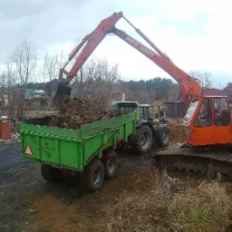 Pomarańczowa koparka gąsienicowa z łyżką załadowuje ziemię do zielonej przyczepy ciągniętej przez traktor na terenie budowy z widocznymi domami w tle.