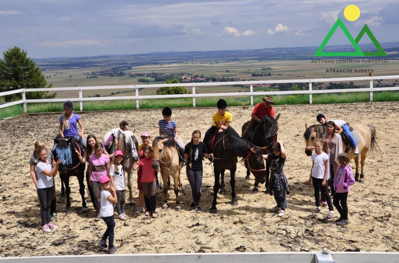 Grupa dzieci i dorosłych, część na koniach, pozuje do zdjęcia na piaszczystym padoku z widokiem na krajobraz i logo 'Aktywne Centrum Turystyki Atrakcje Sudety' w tle.