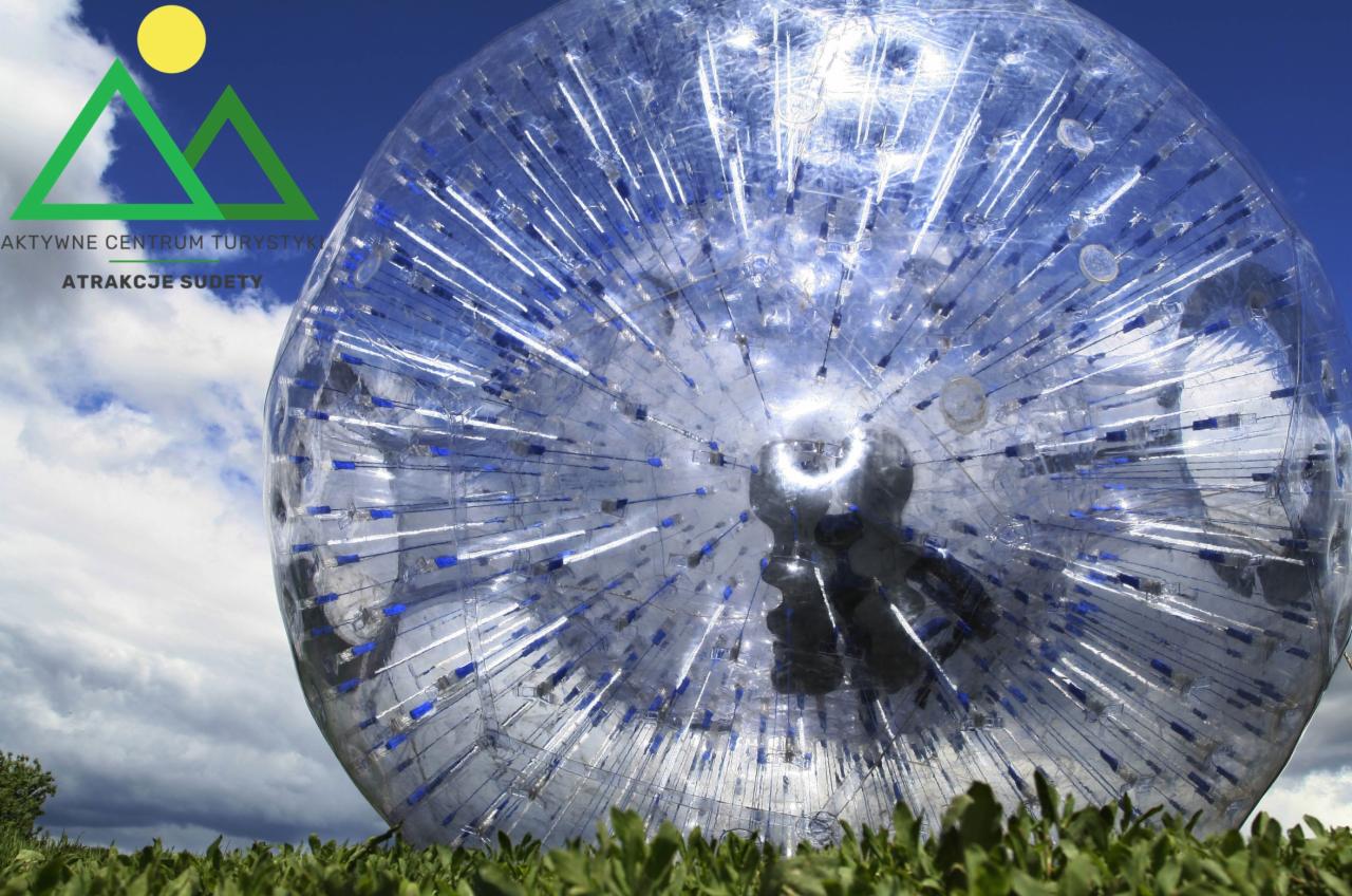 Przezroczysta, duża kula zorbingowa z niebieskimi elementami, leżąca na zielonej trawie, na tle błękitnego nieba z chmurami. Wewnątrz kuli widoczne sylwetki osób.