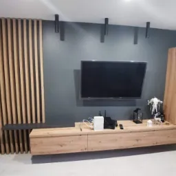 Nowoczesny salon z drewnianą ścianką działową, szafką RTV w odcieniach drewna i szarą ścianą z oświetleniem punktowym.