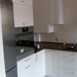 Meble Na Wymiar WNW - Nowoczesna, jasna kuchnia z białymi frontami szafek i ciemnym blatem, widoczny fragment lodówki ze stali nierdzewnej, piekarnik i płyta gazowa, górne szafki wiszące.