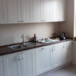 Meble Na Wymiar WNW - Jasna kuchnia z białymi szafkami i ciemnym blatem, widok na zlew, baterię i akcesoria kuchenne.