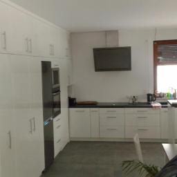 Meble Na Wymiar WNW - Jasna kuchnia z białymi, lakierowanymi frontami szafek, zabudowaną lodówką i piekarnikiem, czarnym blatem, oknem z drewnianymi żaluzjami i lampami wiszącymi na długich kablach.