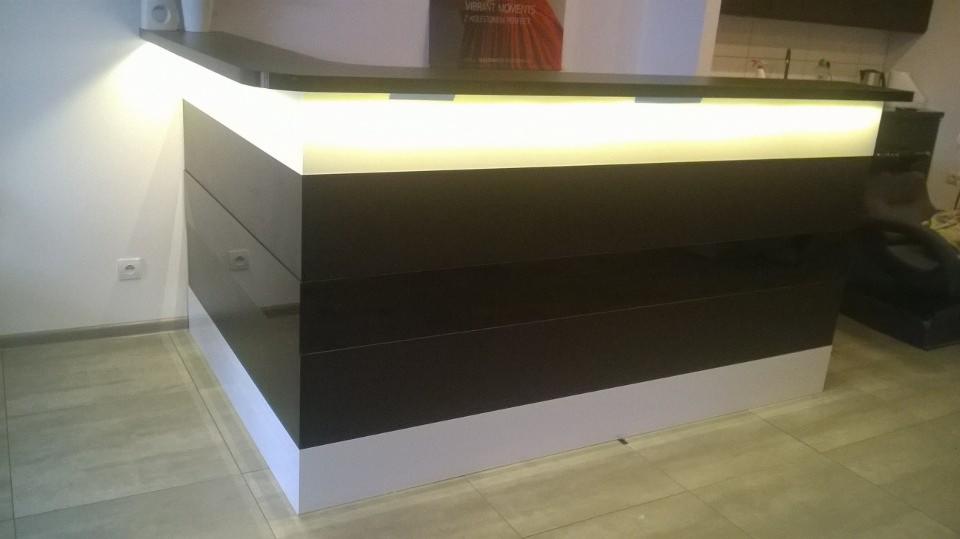 Nowoczesna lada recepcyjna z podświetleniem LED, front w kolorze ciemnego brązu, dolna część w kolorze białym, widoczne gniazdko elektryczne na ścianie w tle.