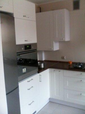 Nowoczesna, jasna kuchnia z białymi frontami szafek i ciemnym blatem, widoczny fragment lodówki ze stali nierdzewnej, piekarnik i płyta gazowa, górne szafki wiszące.