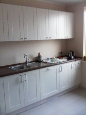 Jasna kuchnia z białymi szafkami i ciemnym blatem, widok na zlew, baterię i akcesoria kuchenne.