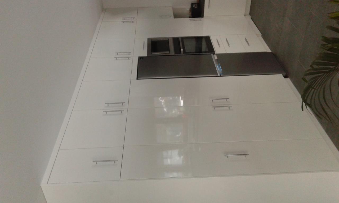 Wysoka zabudowa kuchenna z białymi, lakierowanymi frontami, zawierająca wbudowany piekarnik i lodówkę, z subtelnymi, podłużnymi uchwytami.