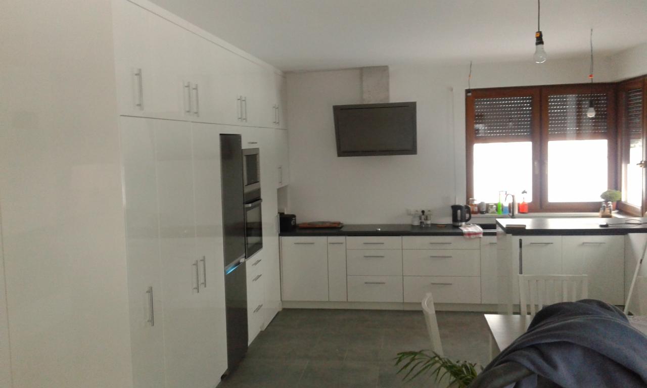 Jasna kuchnia z białymi, lakierowanymi frontami szafek, zabudowaną lodówką i piekarnikiem, czarnym blatem, oknem z drewnianymi żaluzjami i lampami wiszącymi na długich kablach.