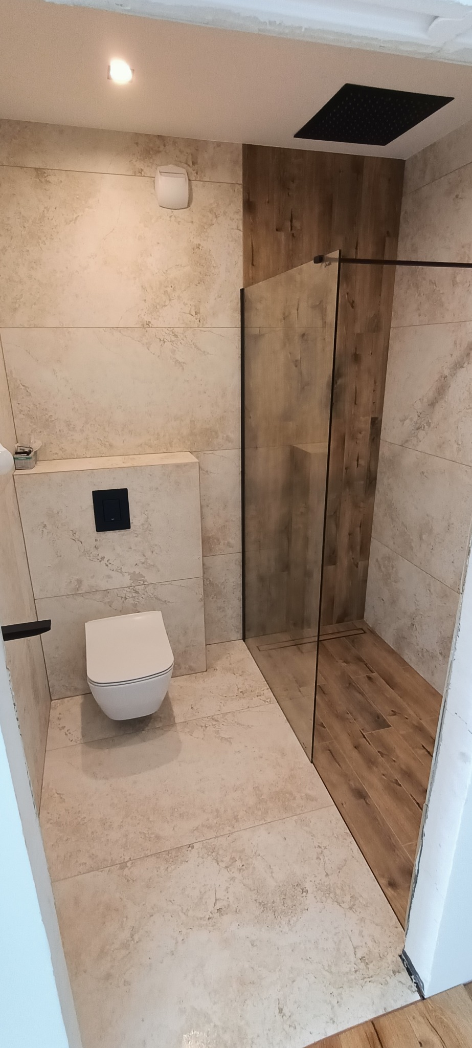 Elegancka łazienka po remoncie: beżowe płytki imitujące kamień, kabina prysznicowa ze szklaną ścianką i deszczownicą w kolorze czarnym, wisząca toaleta.