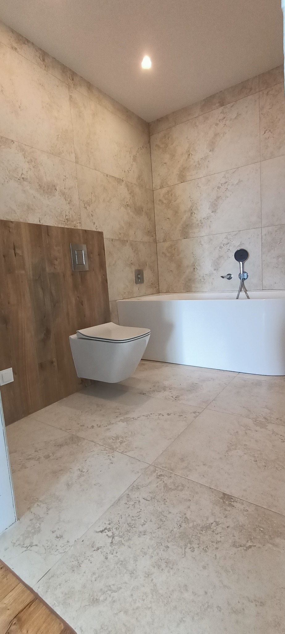 Nowoczesna łazienka z beżowymi płytkami imitującymi marmur, podwieszana toaleta i biała wanna. Minimalistyczny design z drewnianym akcentem za toaletą.