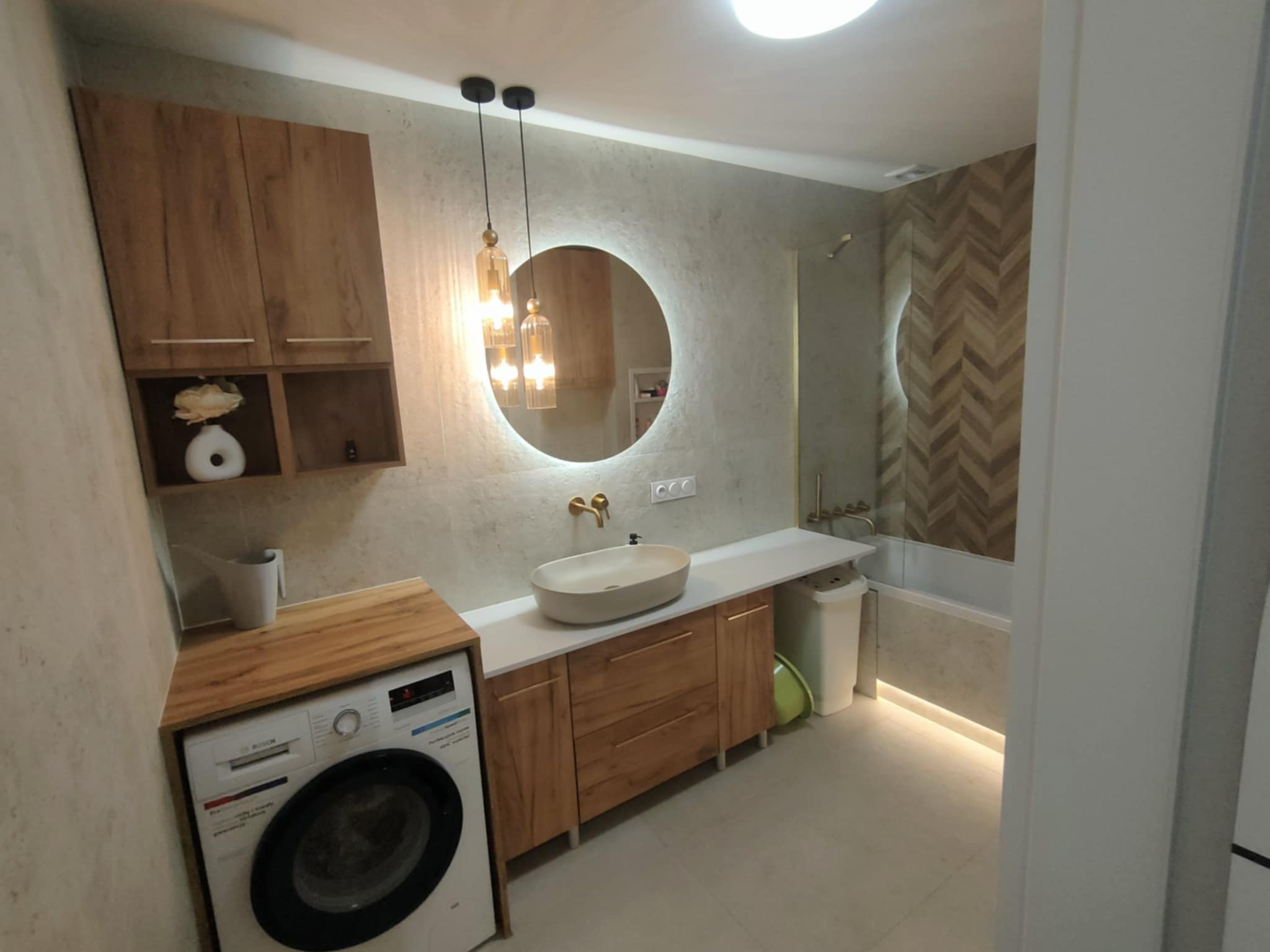 Nowoczesna łazienka z okrągłym lustrem podświetlanym LED, drewnianymi szafkami i pralką. Uwagę przykuwają wiszące lampy i dekoracyjna ściana z motywem jodełki.