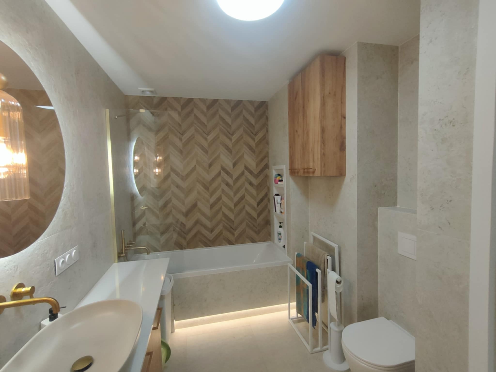 Nowoczesna łazienka z okrągłym lustrem z podświetleniem, drewnianą szafką, wanną z mozaiką w jodełkę i białym blatem z umywalką.