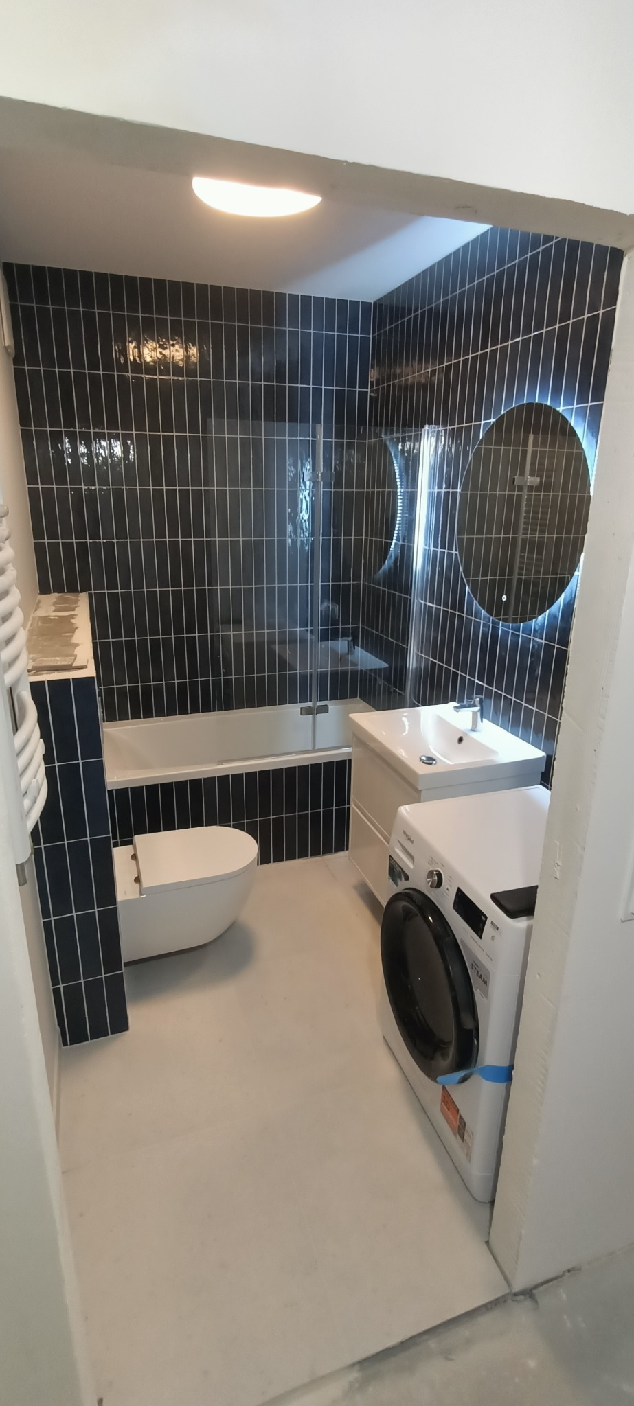 Wykończona łazienka z czarnymi, pionowo ułożonymi płytkami, okrągłym lustrem z podświetleniem LED, białą armaturą i pralką. Nowoczesny design.