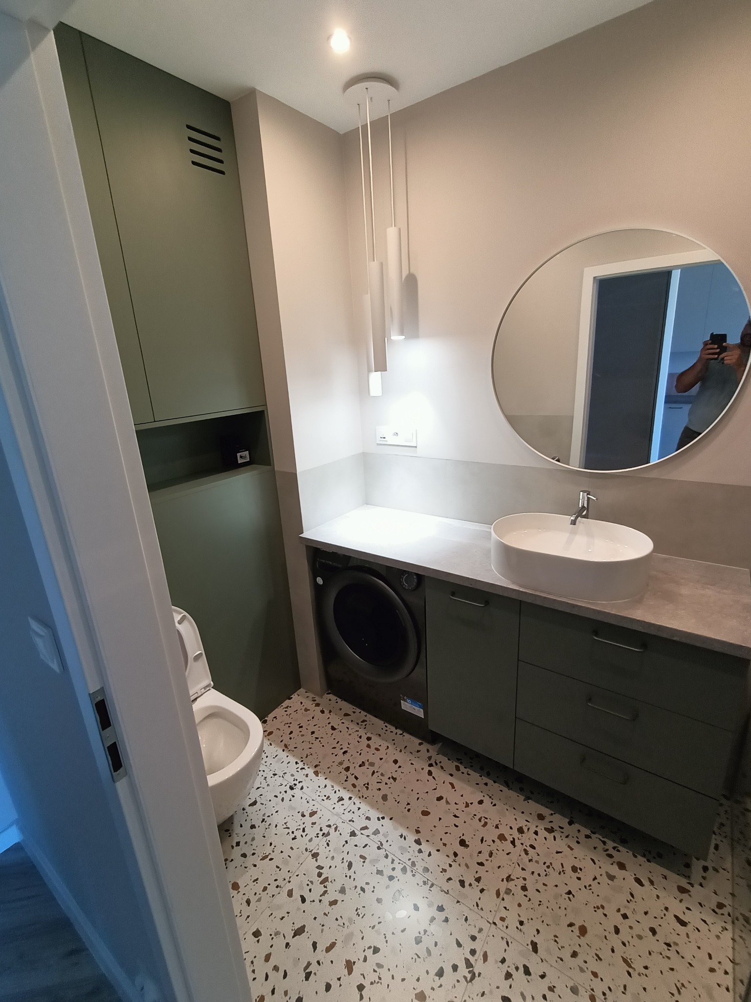 Nowoczesna łazienka z umywalką nablatową, szafkami w odcieniach zieleni i podłogą lastryko. Okrągłe lustro odbija fragment sylwetki osoby robiącej zdjęcie.