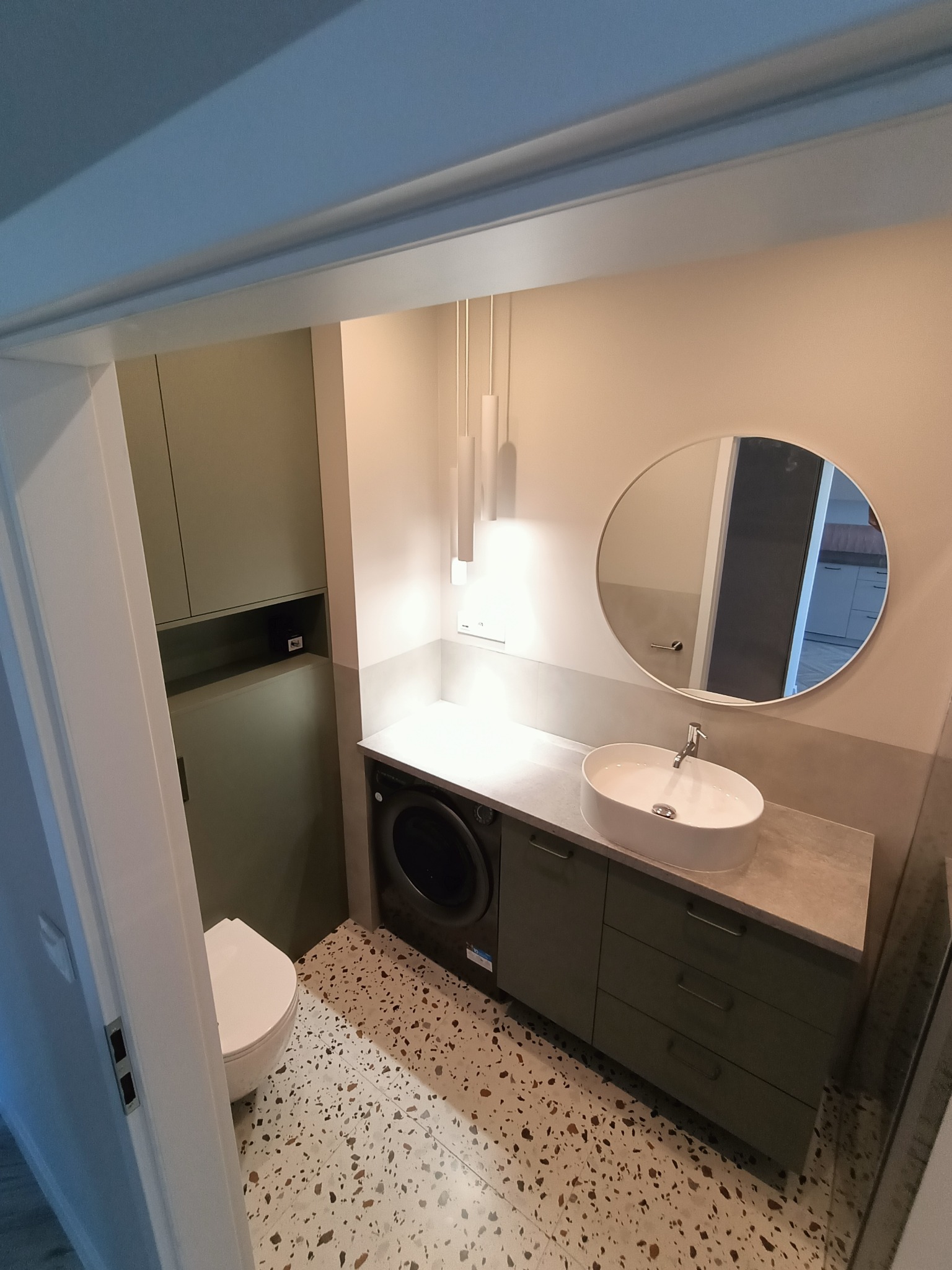 Elegancka łazienka z owalnym lustrem, umywalką nablatową i szafkami w odcieniach zieleni. Na podłodze lastryko. W tle widoczna pralka.