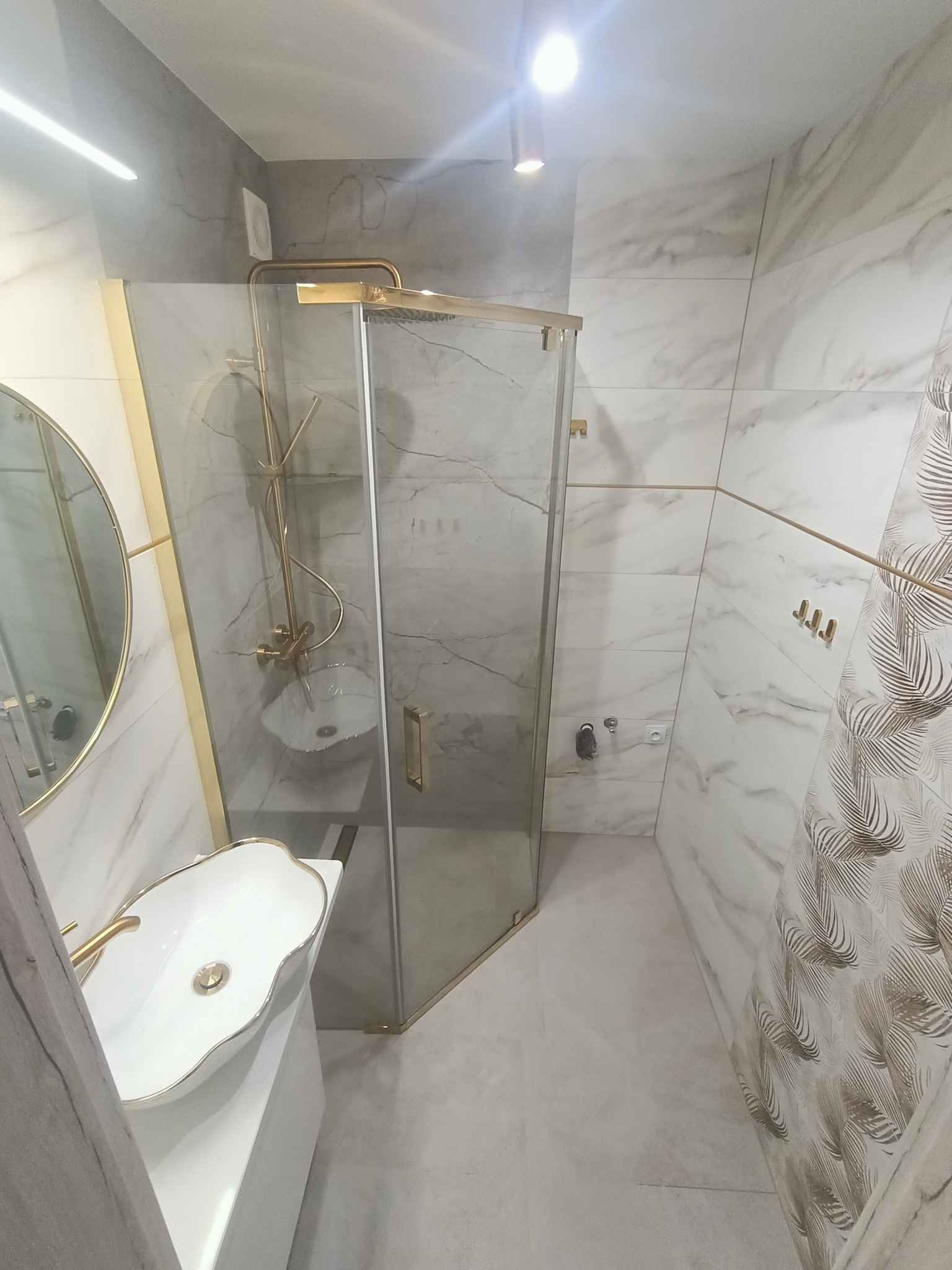 Elegancka łazienka po remoncie: kabina prysznicowa ze złotymi detalami, umywalka na szafce i płytki imitujące marmur z motywem liści. Nowoczesny design.