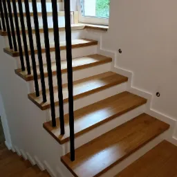 Nowoczesne schody z drewnianymi stopniami i czarnymi, pionowymi balustradami, oświetlone naturalnym światłem z okna na podeście.