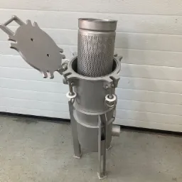 Metalowy filtr cylindryczny z perforowaną wkładką, częściowo otwartą pokrywą i mocowaniami na śruby, stojący na betonowej podłodze przed białą bramą garażową.