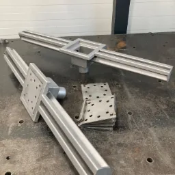 Dwa metalowe elementy konstrukcyjne z kwadratowymi podstawami i podłużnymi profilami leżące na metalowym stole warsztatowym, obok stos metalowych płytek z otworami.