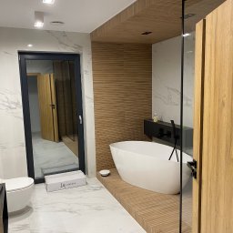 JM STUDIO Jacek Jabłoński, Marcin Pająk - Nowoczesna łazienka z wolnostojącą wanną na podwyższeniu z drewnianych paneli, marmurowymi płytkami i szklaną kabiną prysznicową, widoczna biała ceramika sanitarna i pudełko po produkcie.
