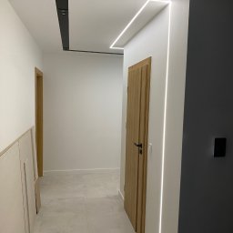 JM STUDIO Jacek Jabłoński, Marcin Pająk - Nowoczesny korytarz z oświetleniem LED wzdłuż sufitu i ścian, drewniane drzwi z czarną klamką, szare płytki podłogowe i panele kartonowo-gipsowe oparte o ścianę po lewej stronie.