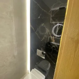 Nowoczesna łazienka z czarnymi marmurowymi płytkami, podświetlanym lustrem i wiszącą toaletą, oświetlona pionową listwą LED.