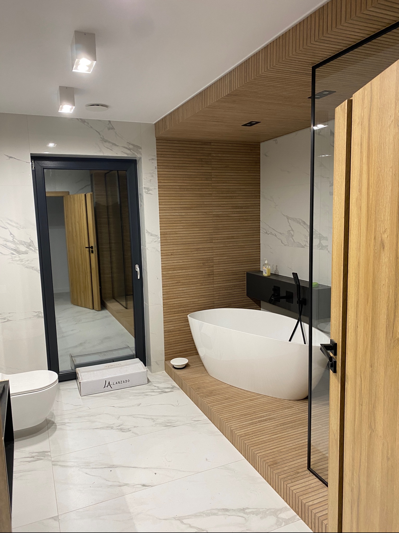 Nowoczesna łazienka z wolnostojącą wanną na podwyższeniu z drewnianych paneli, marmurowymi płytkami i szklaną kabiną prysznicową, widoczna biała ceramika sanitarna i pudełko po produkcie.