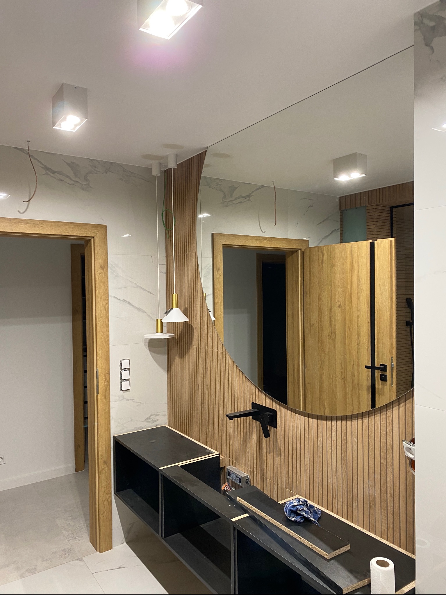 Łazienka w trakcie wykańczania: widoczna zabudowa umywalkowa w czarnym kolorze, lustro w okrągłej ramie z drewnianych listewek, marmurowe płytki na ścianie oraz wisząca lampa z białym kloszem.