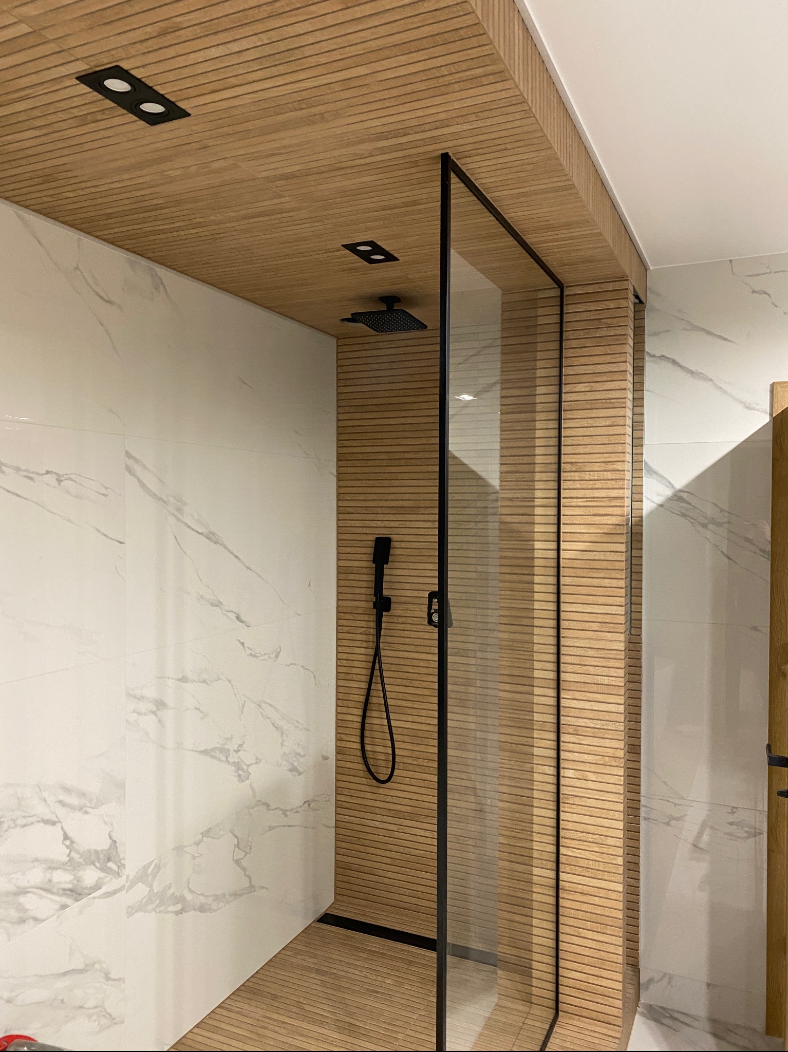 Nowoczesna kabina prysznicowa z czarnymi profilami i deszczownicą, wykończona płytkami imitującymi marmur oraz drewnopodobnymi panelami na ścianach i suficie, podkreślona punktowym oświetleniem.