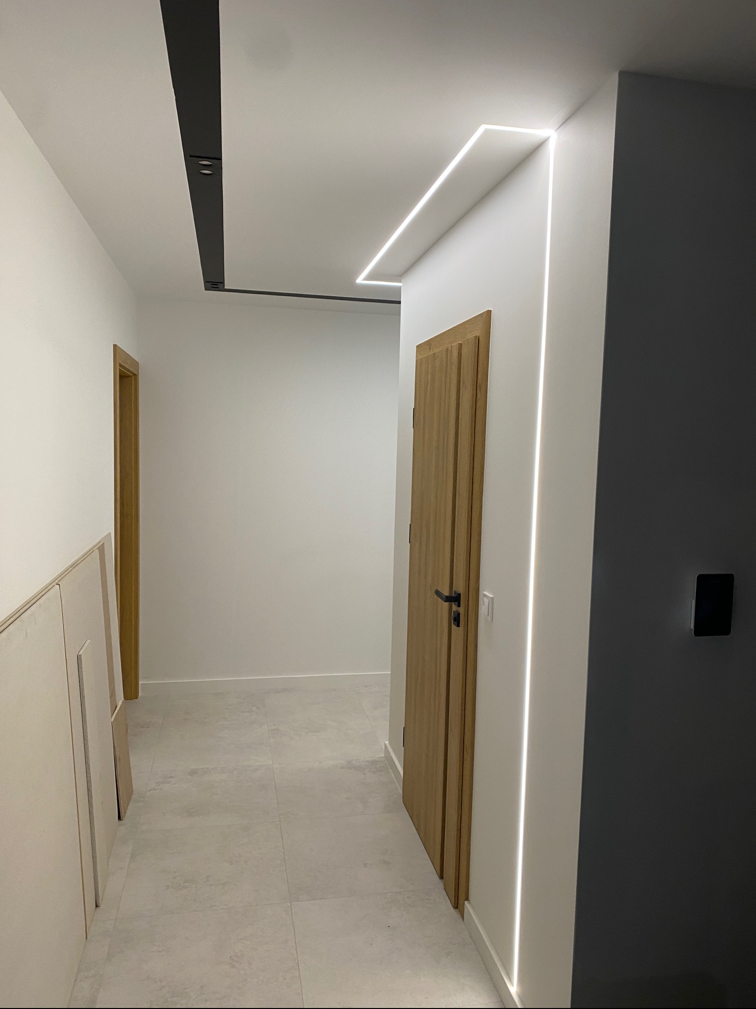 Nowoczesny korytarz z oświetleniem LED wzdłuż sufitu i ścian, drewniane drzwi z czarną klamką, szare płytki podłogowe i panele kartonowo-gipsowe oparte o ścianę po lewej stronie.