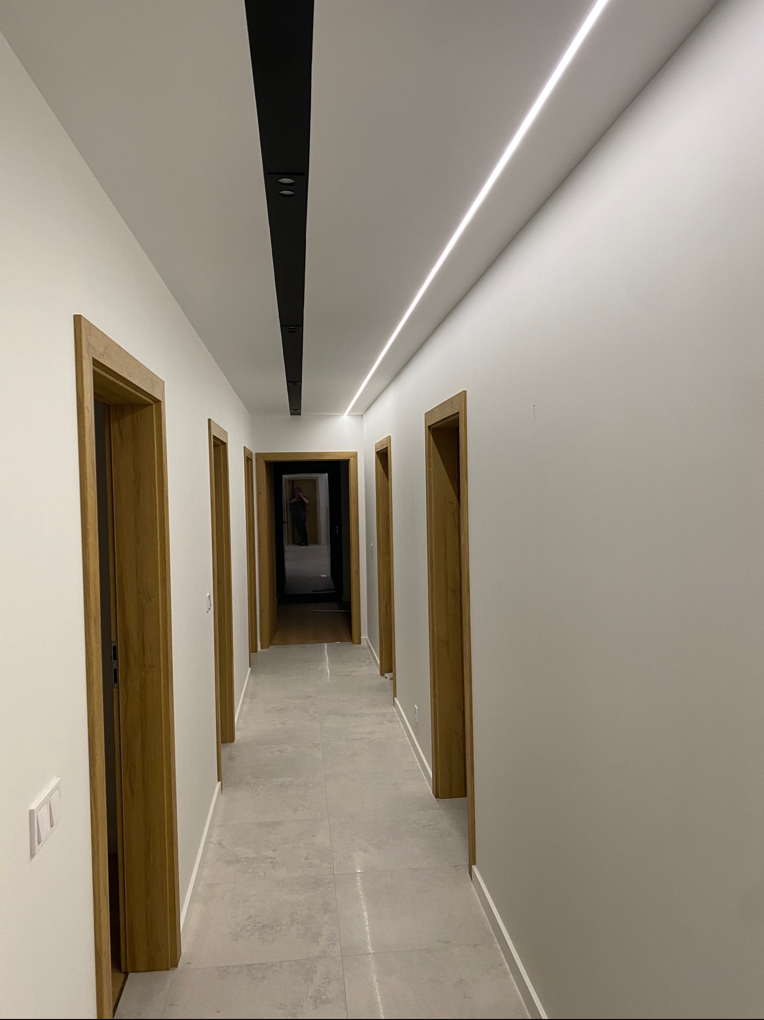Nowoczesny korytarz z białymi ścianami, szarą podłogą z płytek, drewnianymi futrynami drzwi i oświetleniem sufitowym: czarna listwa z reflektorami i liniowa lampa LED.