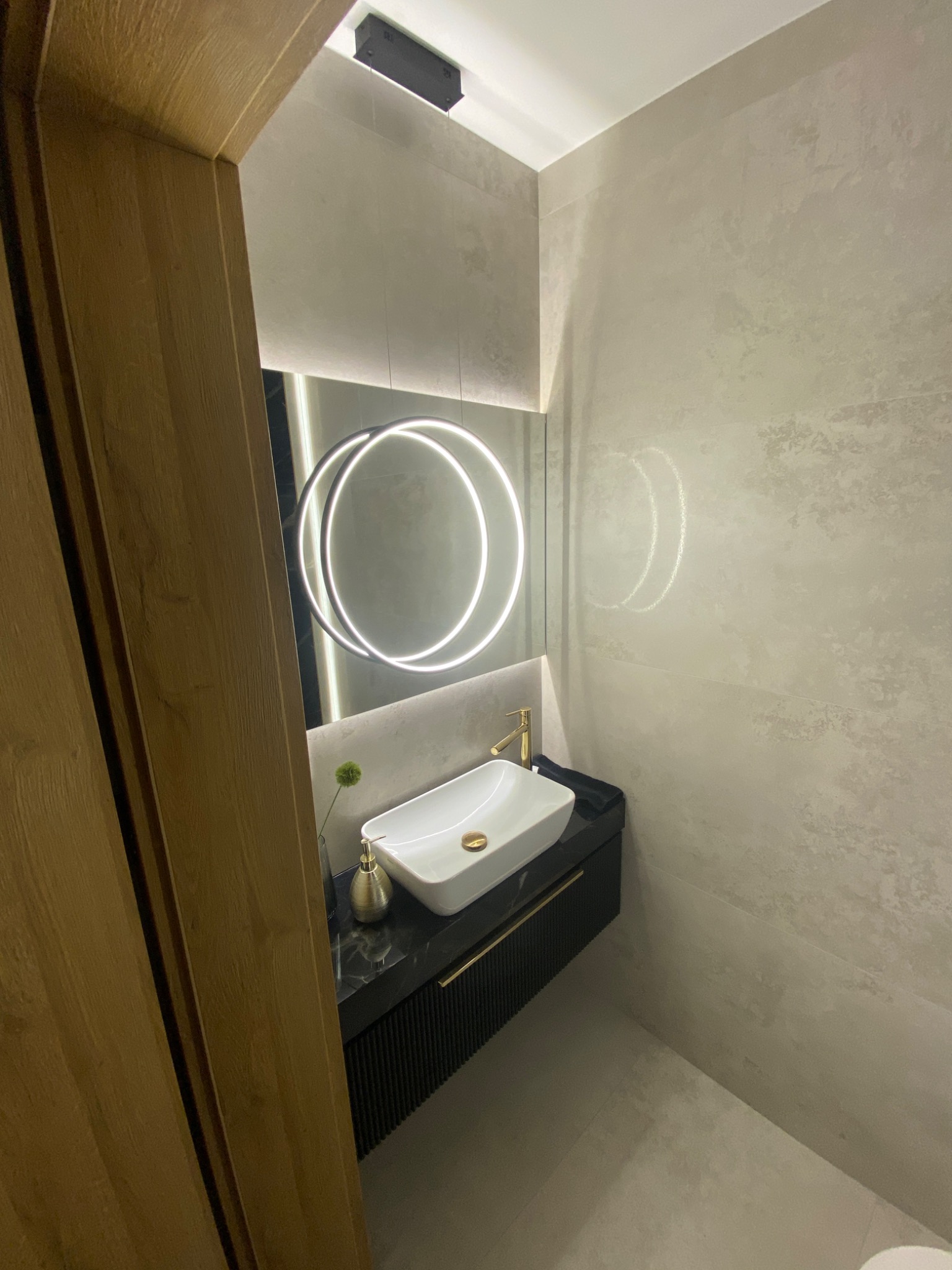 Nowoczesna łazienka w Agatówce z umywalką nablatową, złotą armaturą i okrągłym lustrem z podświetleniem LED, wykończona płytkami w odcieniach szarości.