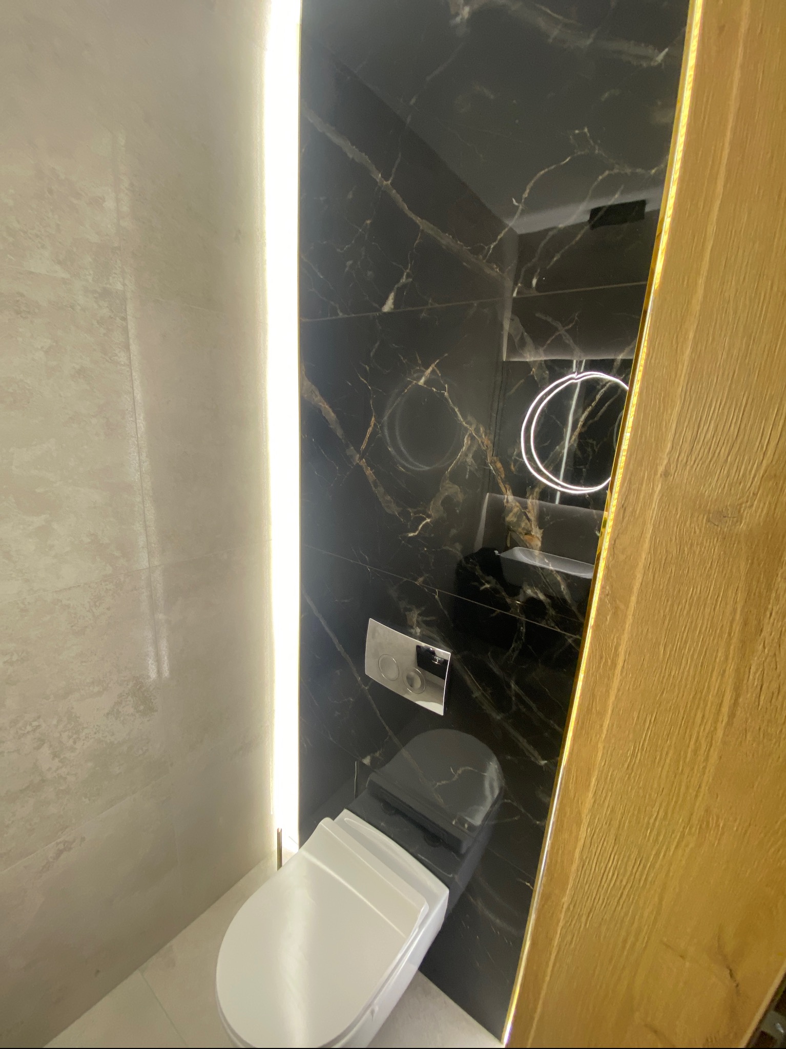 Nowoczesna łazienka z czarnymi marmurowymi płytkami, podświetlanym lustrem i wiszącą toaletą, oświetlona pionową listwą LED.