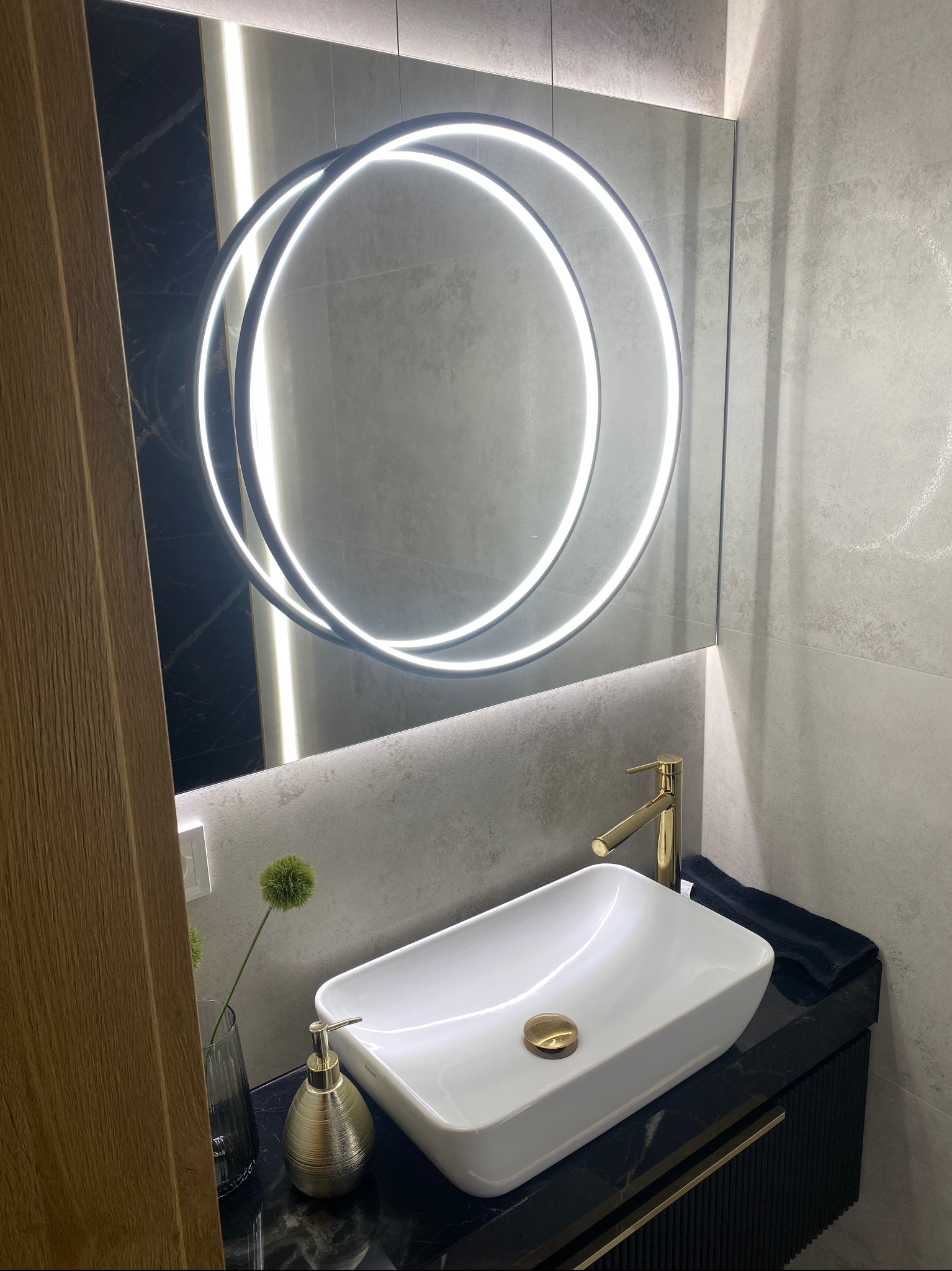 Nowoczesna łazienka z umywalką nablatową, złotą armaturą, podświetlanym okrągłym lustrem i czarnym blatem z marmurowym wzorem.