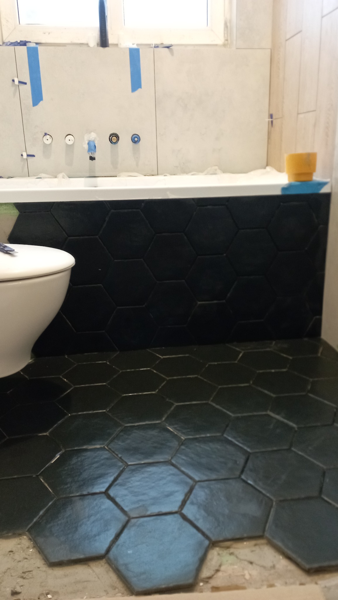 Układanie czarnych, heksagonalnych płytek podłogowych i na obudowie wanny w łazience, widoczne przygotowane przyłącza do baterii, fragment toalety i niedokończone fugowanie.