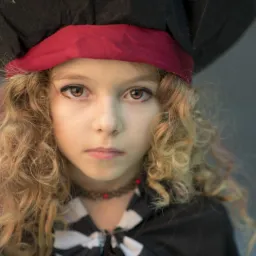 Portret dziewczynki z kręconymi włosami w pirackim kapeluszu i stroju, z delikatnym makijażem oczu.
