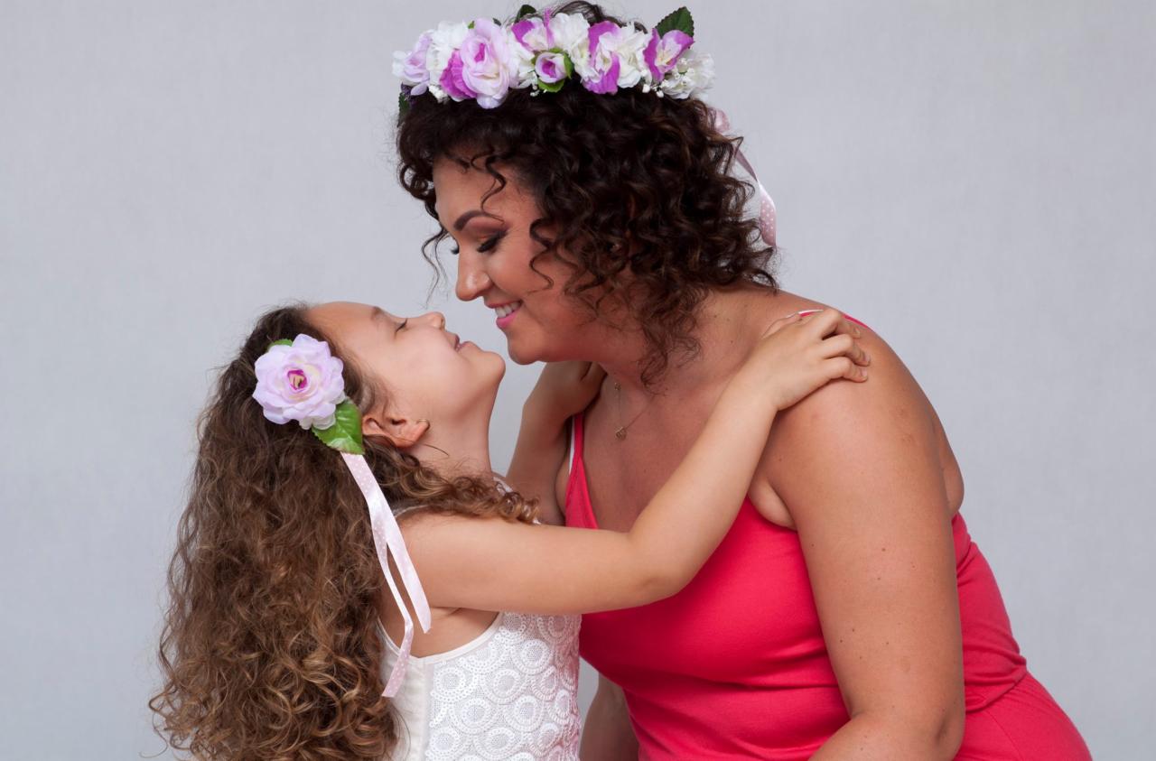 Córka obejmuje uśmiechniętą mamę, obie w kwiatowych wiankach, na jasnym tle.