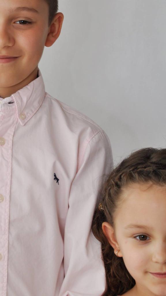 Portret rodzeństwa z Pasłęka: chłopiec w różowej koszuli z logo konika i dziewczynka z warkoczami, oboje częściowo widoczni na neutralnym tle.