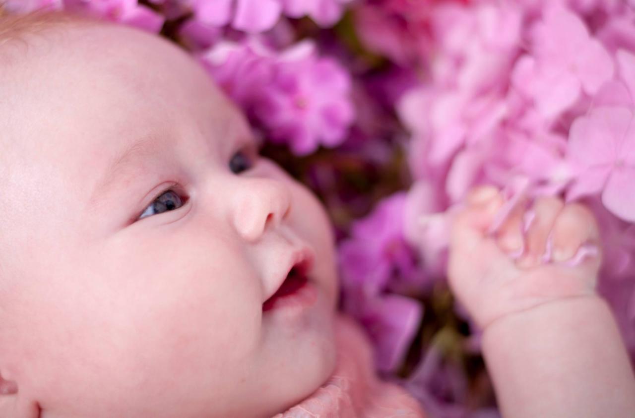 Niemowlę leżące wśród różowych kwiatów hortensji, z otwartymi ustami i delikatnym uśmiechem, patrzy w lewo, mała rączka zaciśnięta w piąstkę.