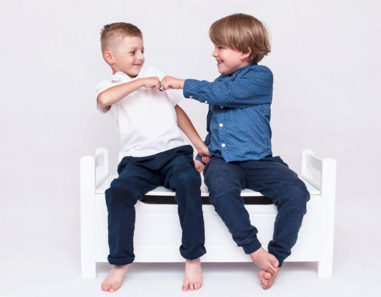 Dwóch uśmiechniętych chłopców siedzi na białej ławeczce, dotykając się pięściami w geście przyjaźni na tle jasnego, jednolitego tła.