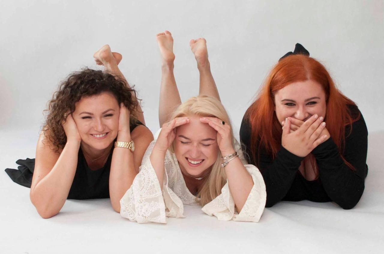 Trzy uśmiechnięte kobiety leżące obok siebie na białym tle, w różnych stylizacjach, z uniesionymi stopami środkowej osoby.