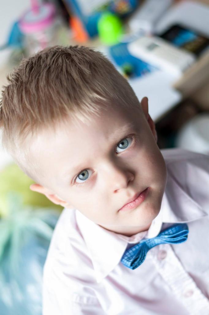 Portret blond chłopca z niebieskimi oczami, ubranego w jasną koszulę i błękitną muchę, uchwycony z góry na rozmytym tle z zabawkami.