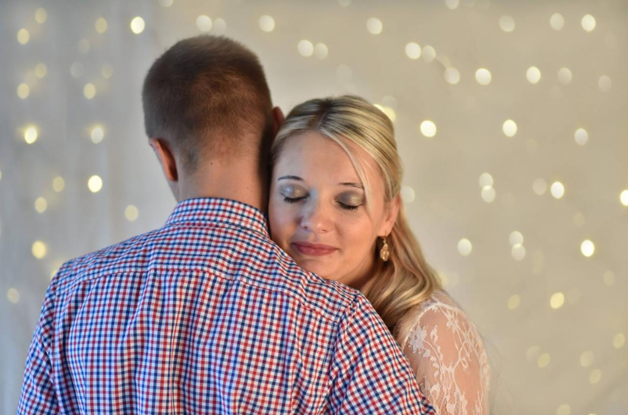 Kobieta z blond włosami i kolczykami, z zamkniętymi oczami, przytula się do mężczyzny w koszuli w czerwoną i niebieską kratę na tle rozświetlonych światełek.