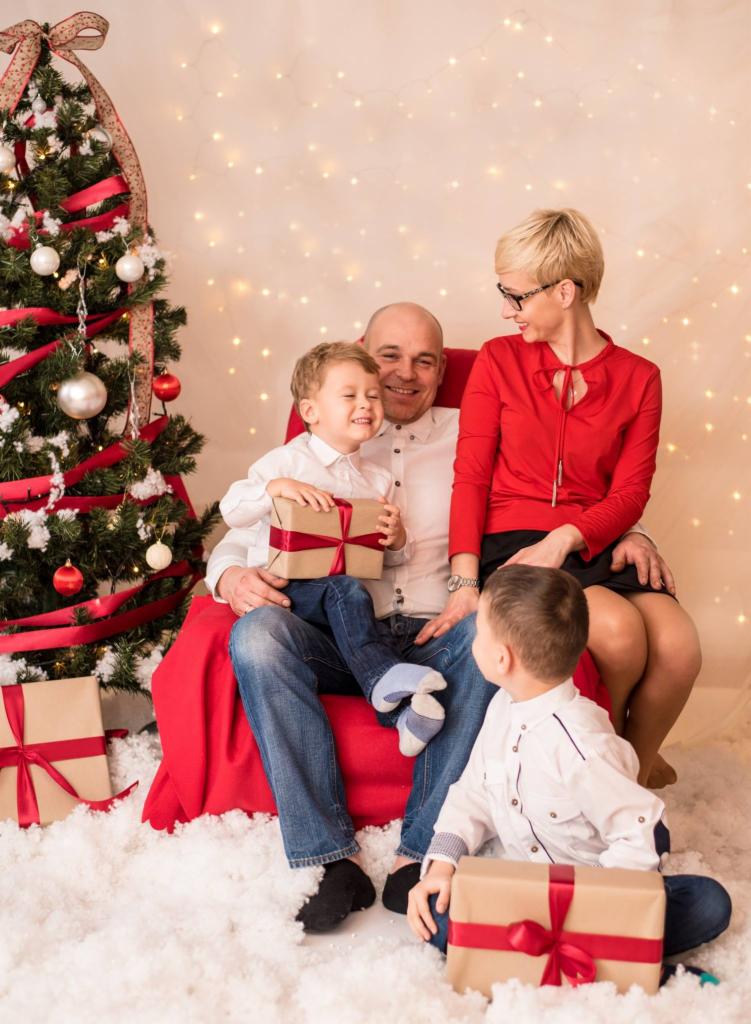 Uśmiechnięta rodzina siedzi na czerwonym fotelu przy choince, mały chłopiec trzyma prezent, a drugi patrzy na niego z dołu, w tle światełka.