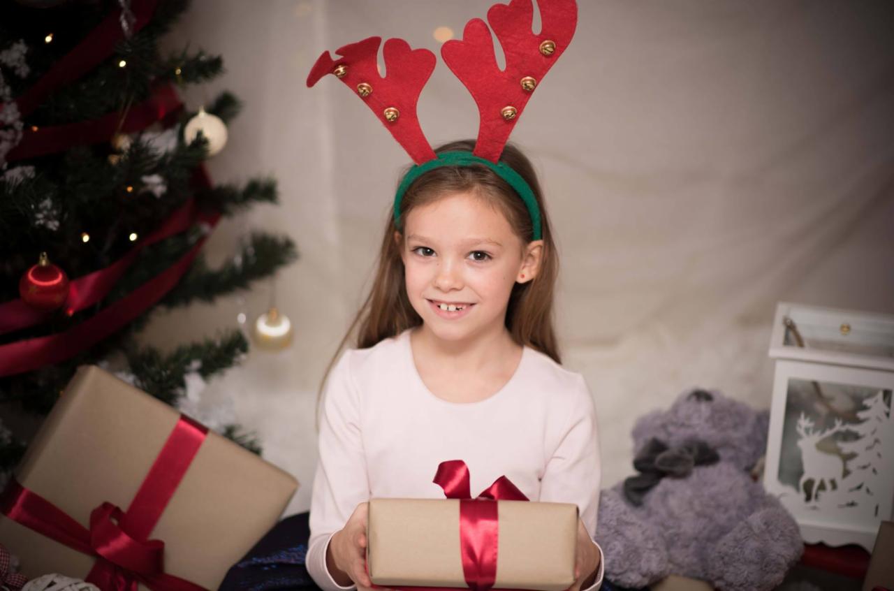 Uśmiechnięta dziewczynka w opasce z rogami renifera trzyma prezent z czerwoną wstążką na tle choinki i pluszowego misia.