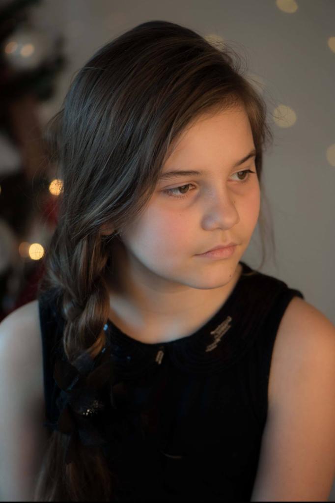 Portret młodej dziewczyny z warkoczem na tle rozmytych światełek choinkowych, ubrana w czarną sukienkę z ozdobnym kołnierzykiem.